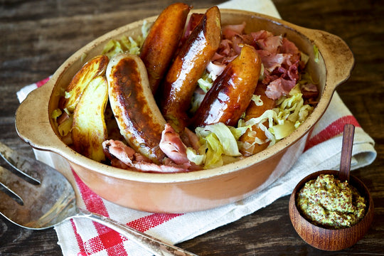 Sausages in a bowl of potatoes and kraut and mustard on the side